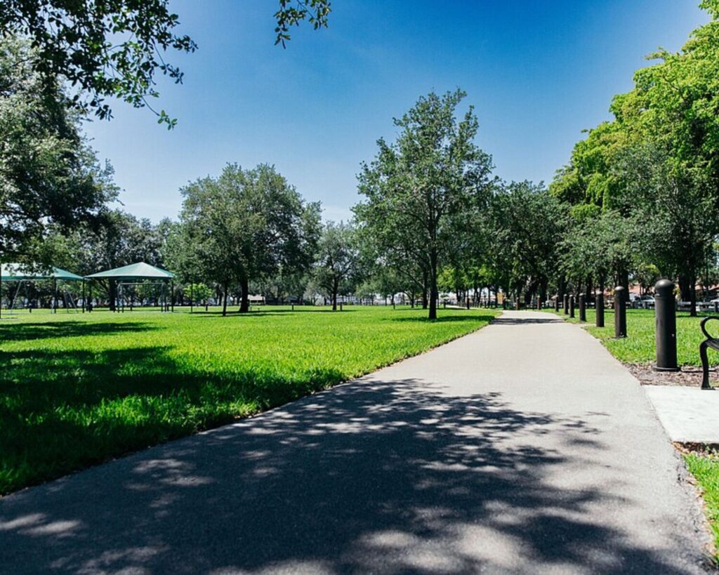 Sunlit park path winding through green lawn and mature trees, with shade canopies in the distance on the left.