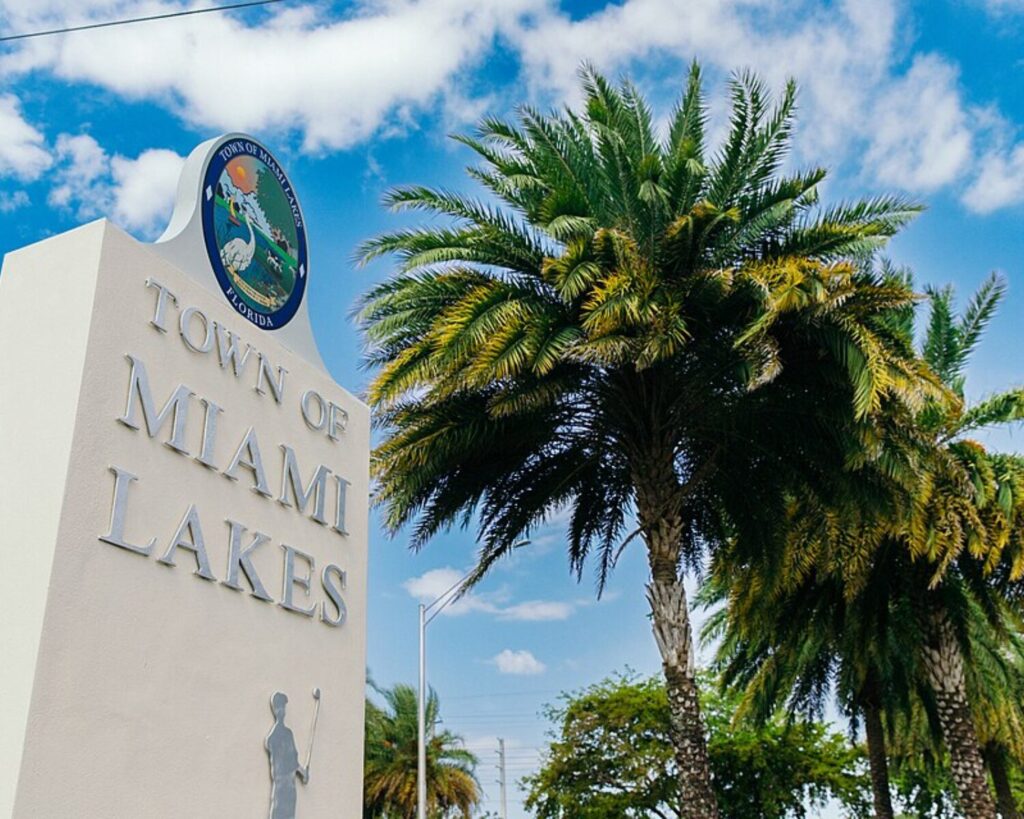 Miami Lakes Drive entrance sign with palm tree, blue sky, and scattered clouds.