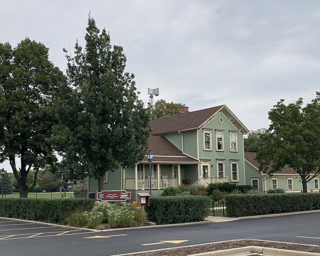 Morton Grove Historical Museum with a green historic house, parking lot, hedges, tall trees, and cloudy sky.