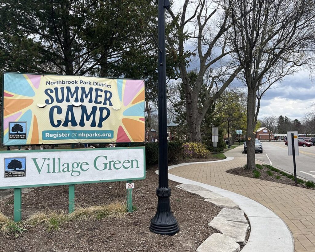 Sign for Northbrook Park District Summer Camp and Village Green, outdoor plaza with brick walkway and trees in spring.