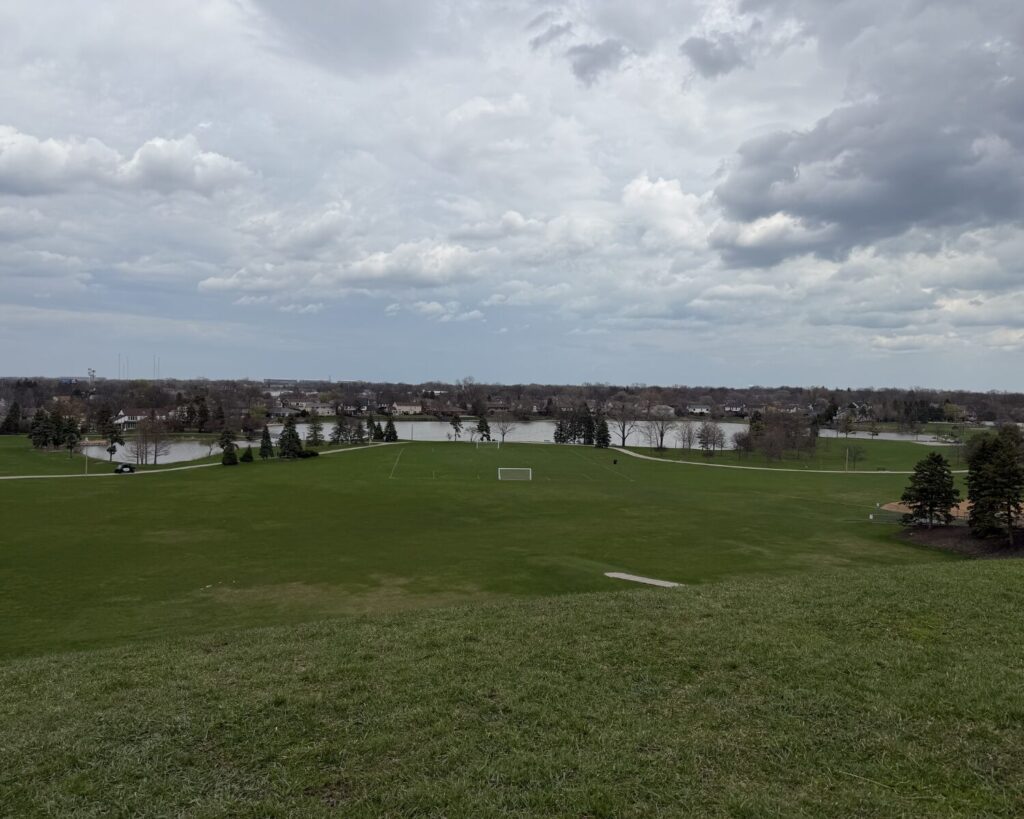 Wide view of a grassy park with a small soccer goal in the center, a lake beyond, and a residential area under a cloudy sky.