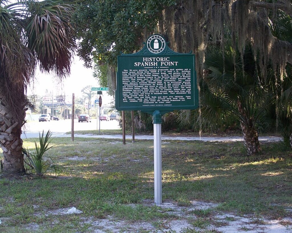 Historic Spanish Point marker sign in Osprey, Florida surrounded by palm trees, grass, and roadside greenery.