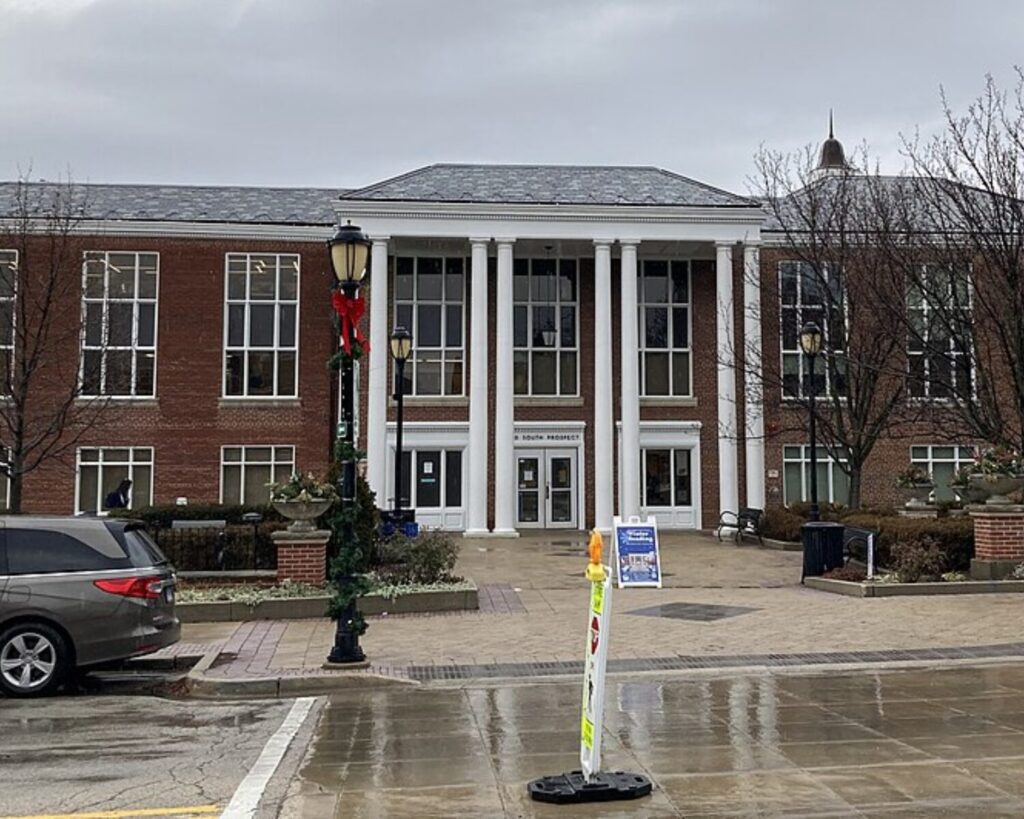 Front view of a brick campus building with a white-columned portico and large windows; holiday wreath on the streetlamp in the foreground, wet pavement.