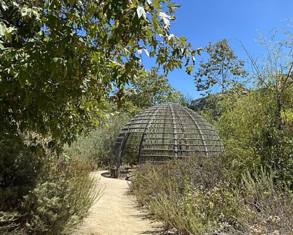 Model of a Tongva kiiy at Ballona Discovery Park with dirt path, native plants, and clear blue sky.
