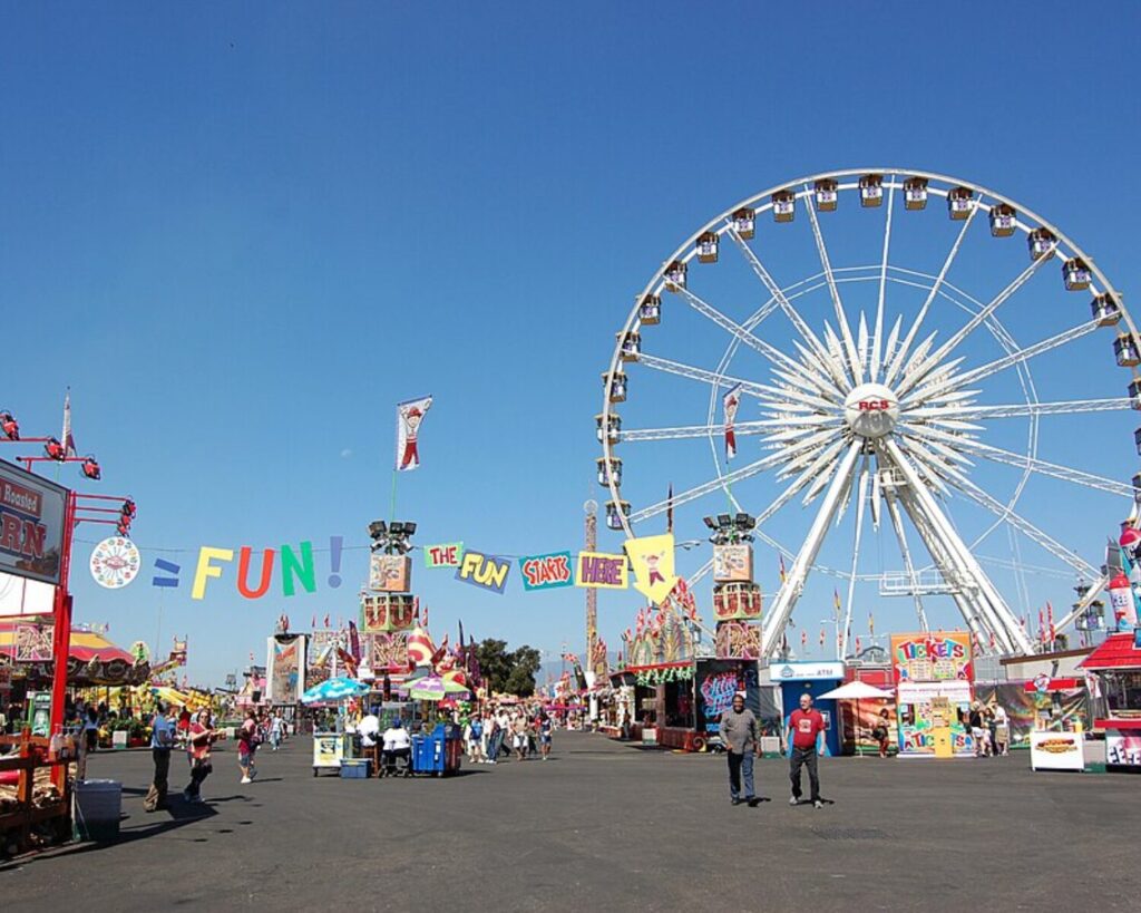 L.A. County Fair with Ferris wheel, food stalls, visitors, and clear blue sky.