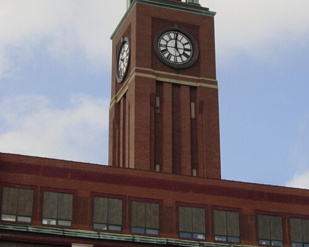 Brick clock tower rising from a red brick building against a blue sky. The clock faces are visible on two sides of the tower.