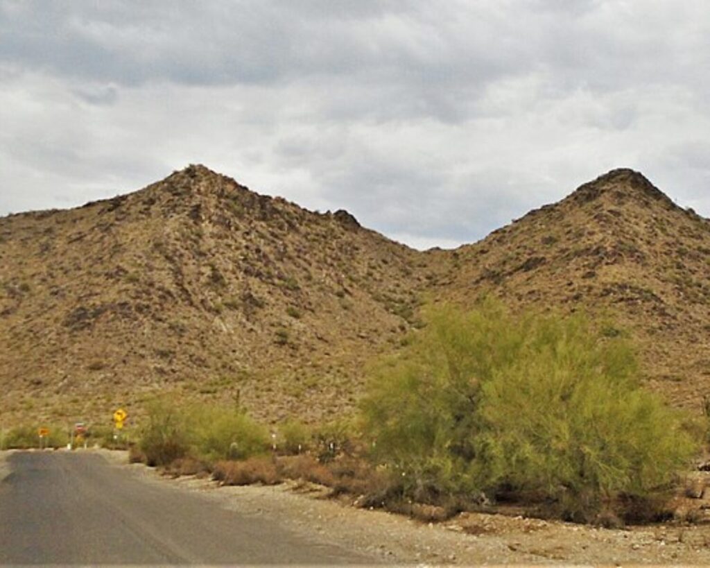 Desert road winds past brown rocky hills under a cloudy sky, with sparse desert shrubs in the foreground.
