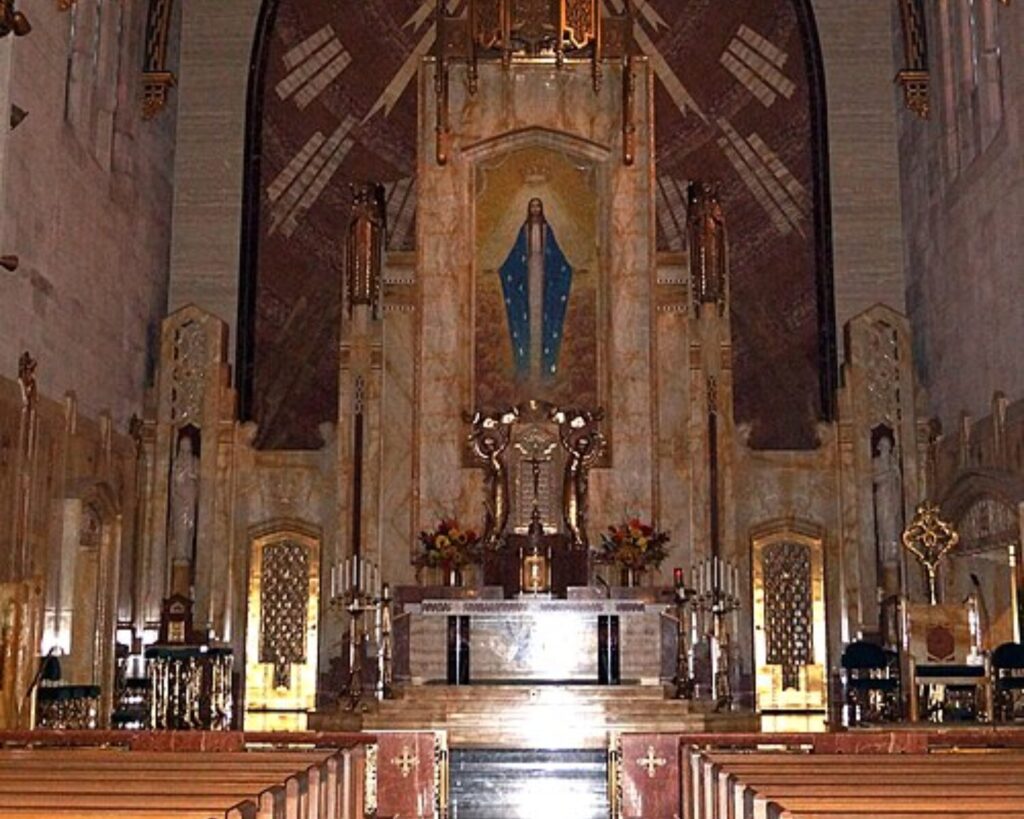 Interior of Queen of All Saints Basilica with a tall arched altar, pews, religious statue, and ornate ceiling.