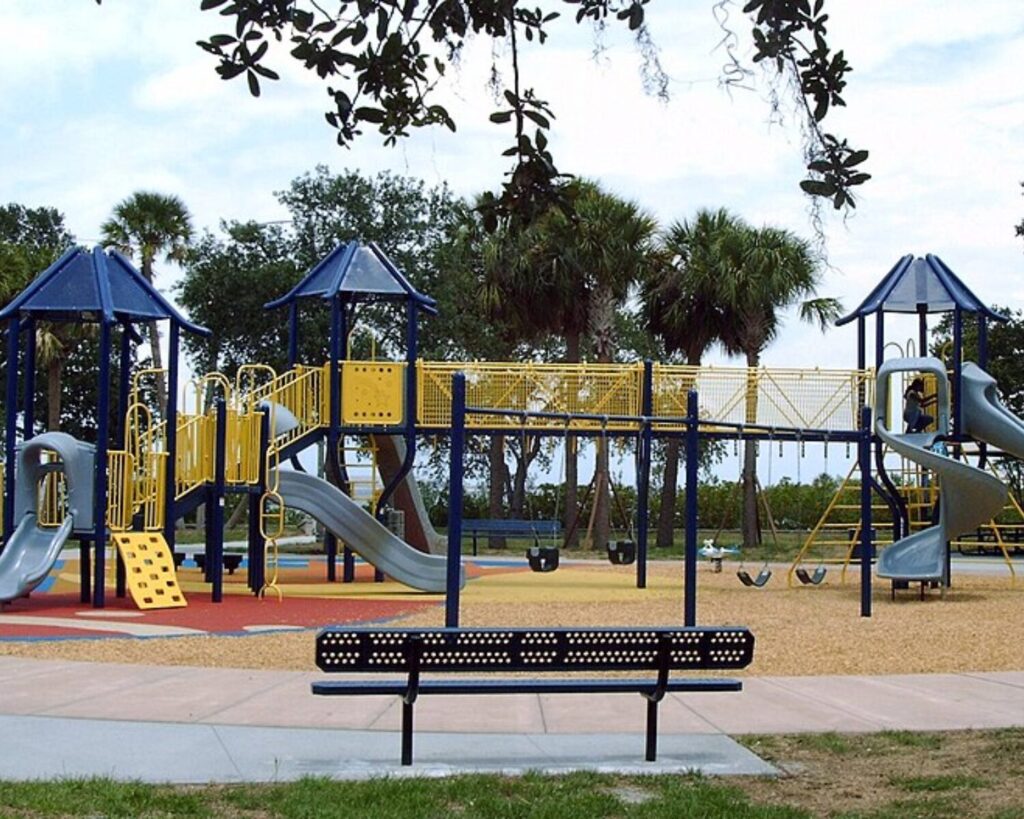 Colorful blue and yellow playground structures with slides and swings in a park, bench in the foreground.