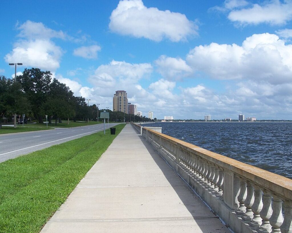 Lakeside sidewalk with railing, grassy verge, and distant apartment buildings under a bright blue sky with clouds.
