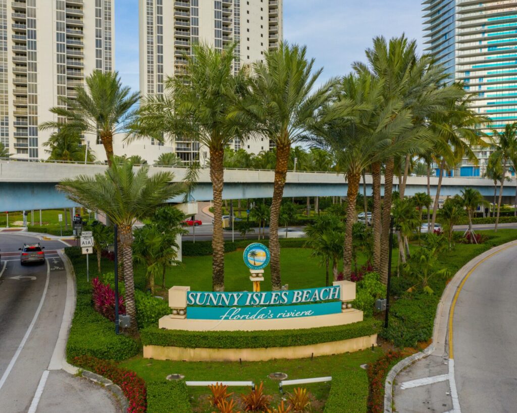Sunny Isles Beach sign with palm trees and tall beachfront buildings in a sunny urban area.