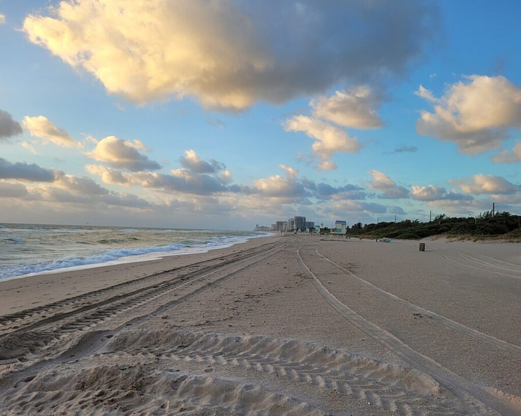 Wide sandy beach with tire tracks along the shoreline, calm waves, and a distant city skyline under a partly cloudy sky in the distance.