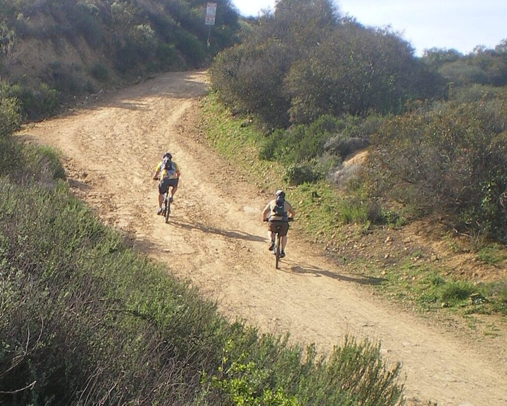 Mountain bikers riding on a dirt trail at Marvin Braude Gateway Park with shrubs and hills.