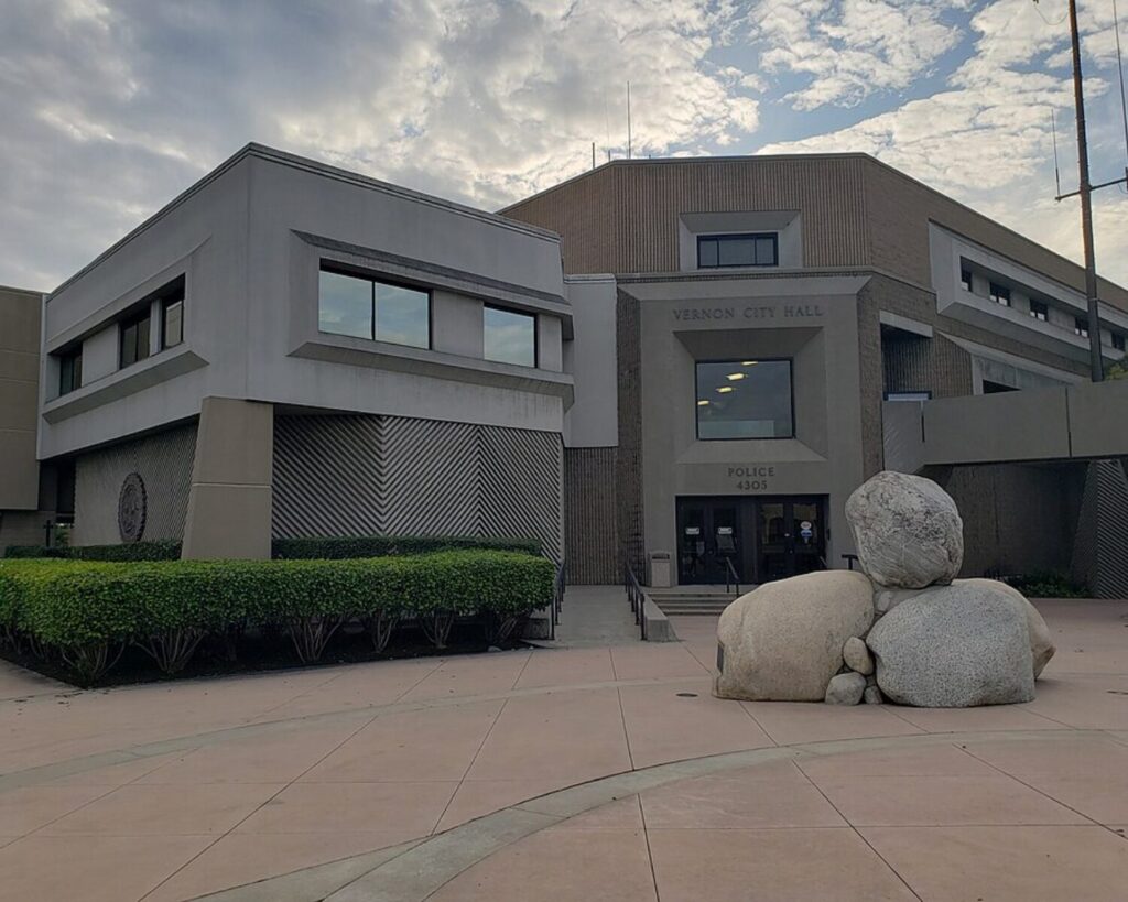 Vernon City Hall building with a front plaza, trimmed hedges, and large decorative rocks under a cloudy sky.