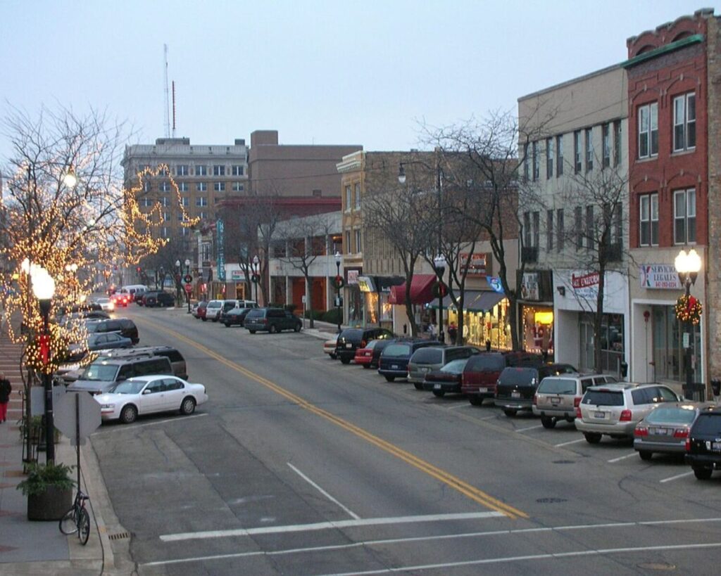 Downtown Waukegan street during the holidays with decorated lights, parked cars, storefronts, and evening sky.
