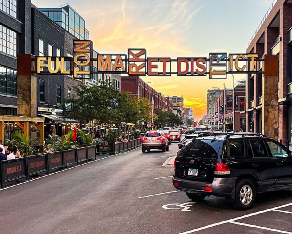 Fulton Market District street in Chicago at sunset with restaurants, traffic, parked cars, and overhead sign.