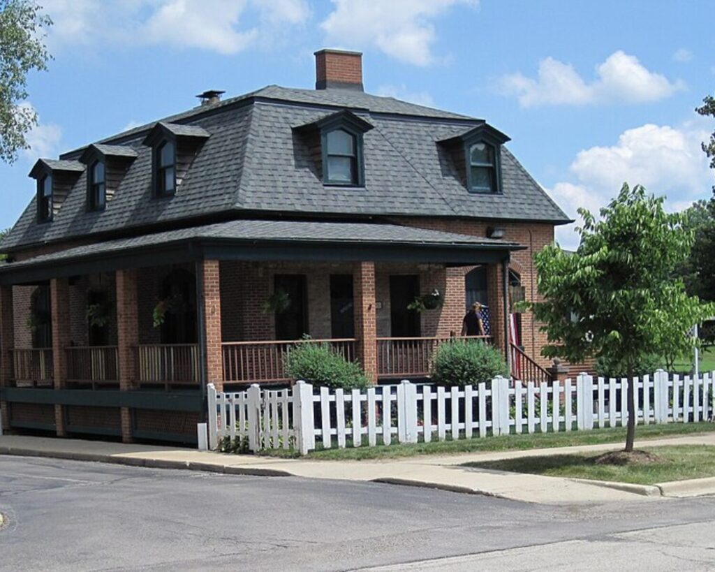 Two-story brick house with a wraparound porch, dormer windows, and a white picket fence on a sunny street.
