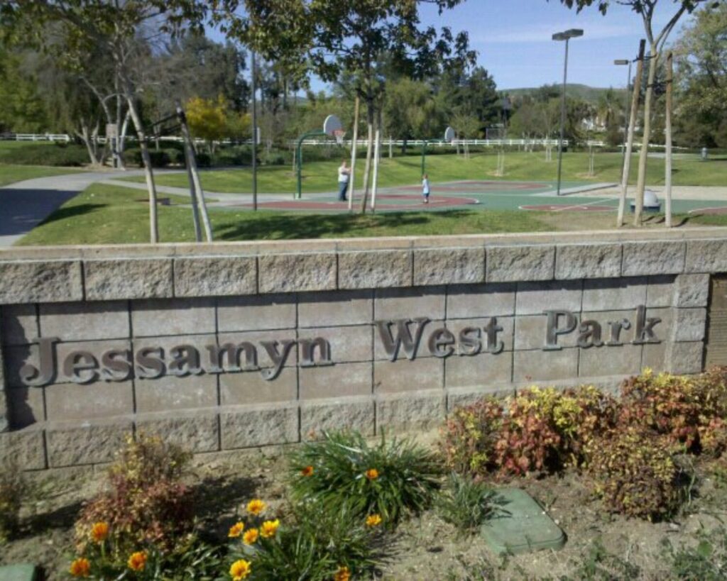 Jessamyn West Park entrance sign with a basketball court, green lawn, trees, and hills in the background.