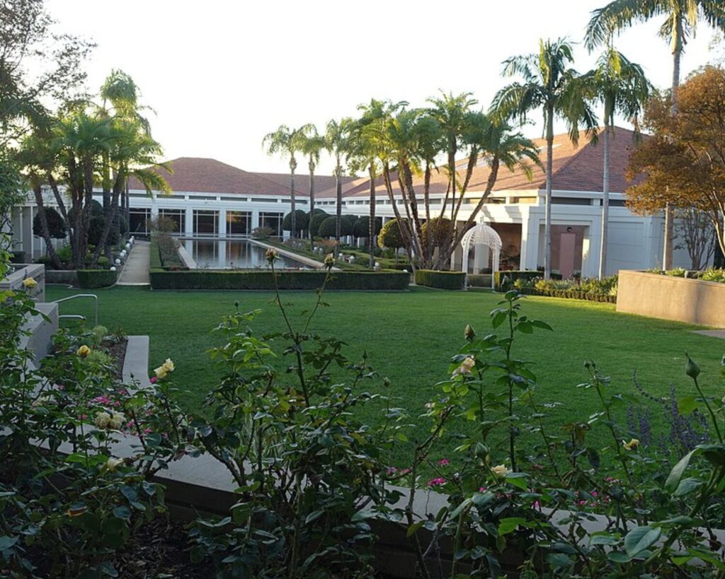 A formal courtyard with a rectangular reflecting pool, trimmed hedges, and a white building lined with glass doors, surrounded by palm trees and green lawn.