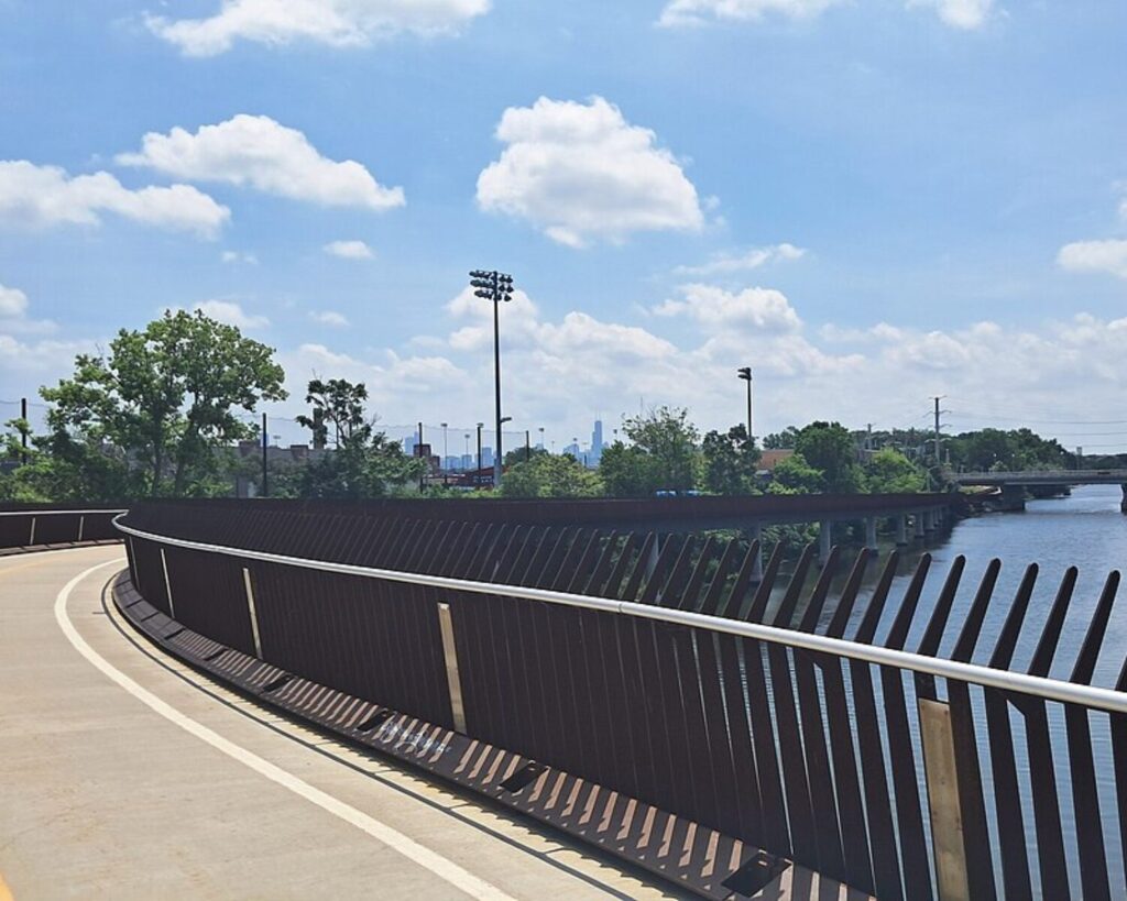 Curved riverside walkway with metal railing, trees, and distant city skyline under a blue, cloud-filled sky.