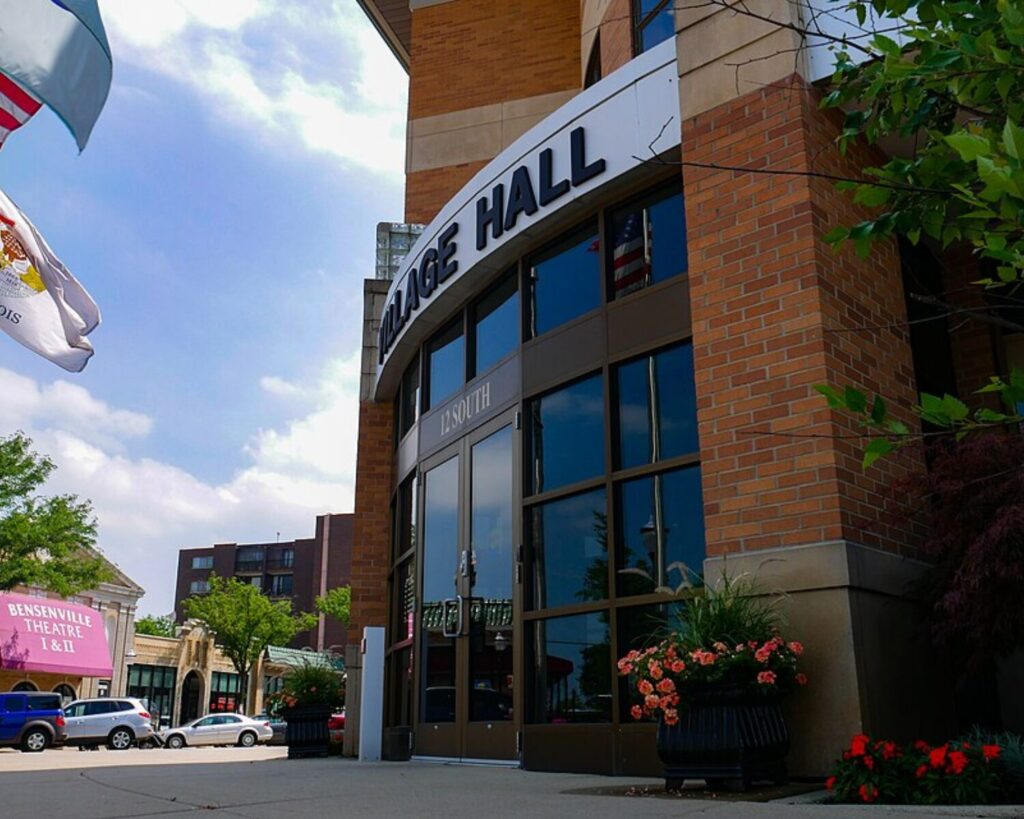 Brick village hall facade with curved white sign reading 'Village Hall' above glass doors, blue sky, and planters with pink flowers nearby, giving a welcoming town-center feel.