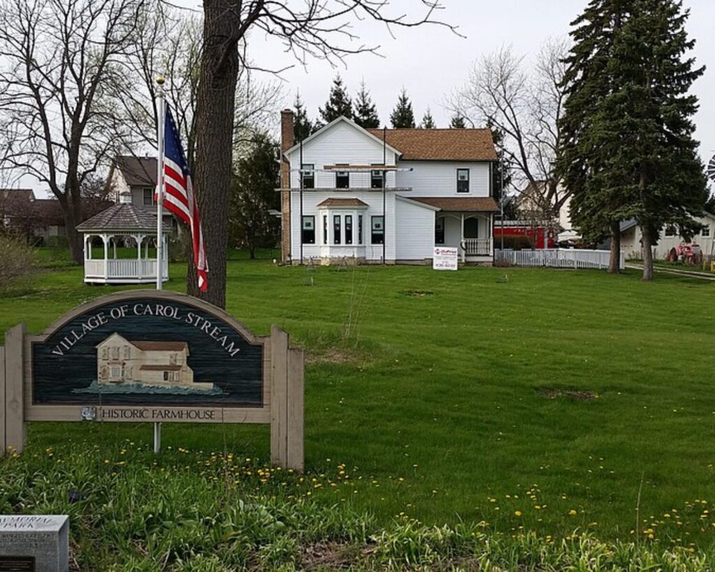 White two-story farmhouse on a grassy lawn with a flagpole, gazebo, and a 'Village of Carol Stream Historic Farmhouse' sign in the foreground.