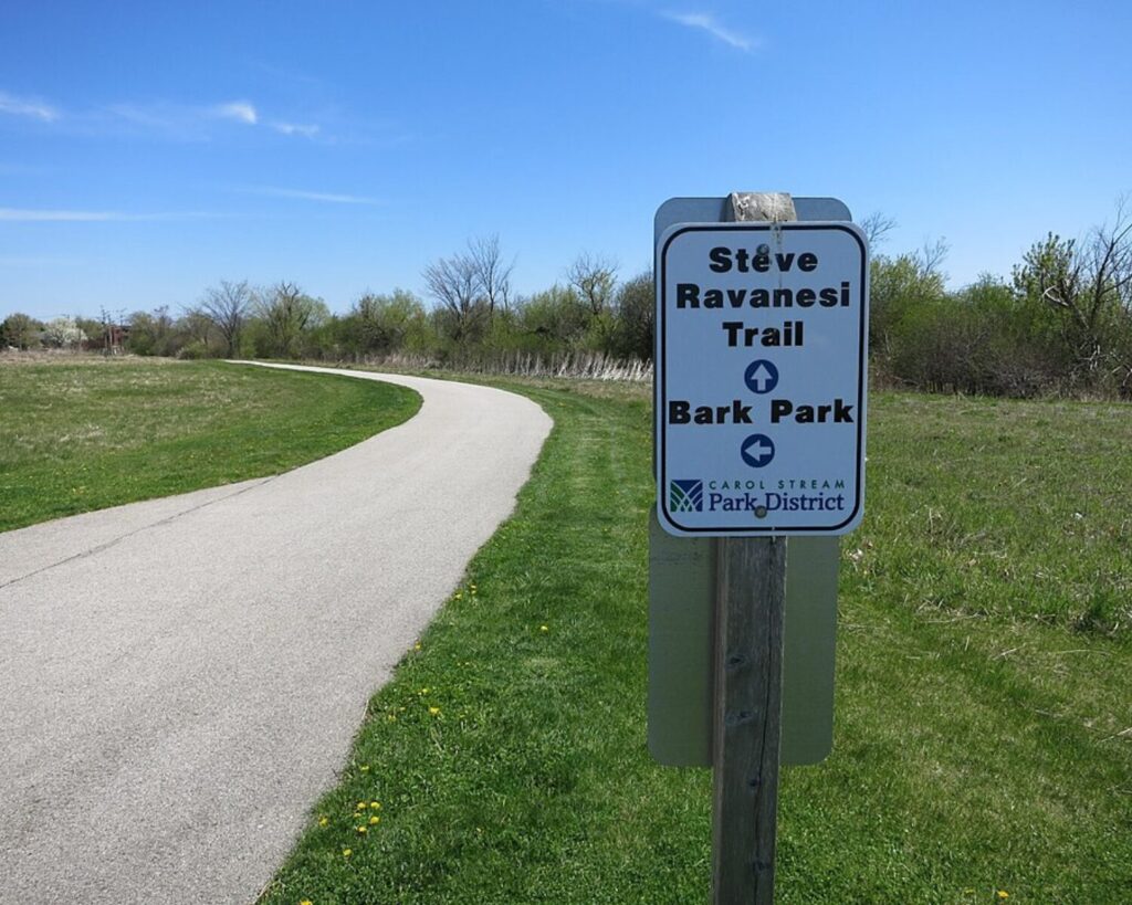 Trail sign on a post reading 'Steve Ravanesi Trail' and 'Bark Park' with arrows, along a paved path in a park on a sunny day
