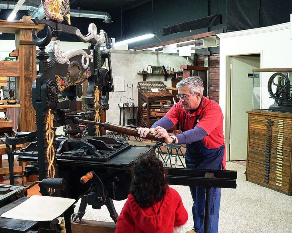 Museum demonstration of an antique printing press with people operating the machine indoors.