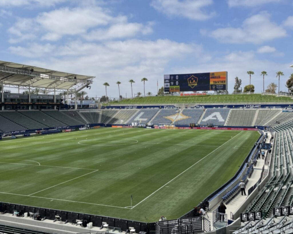 Soccer stadium with green field, empty seats, scoreboard, palm trees, and partly cloudy sky.