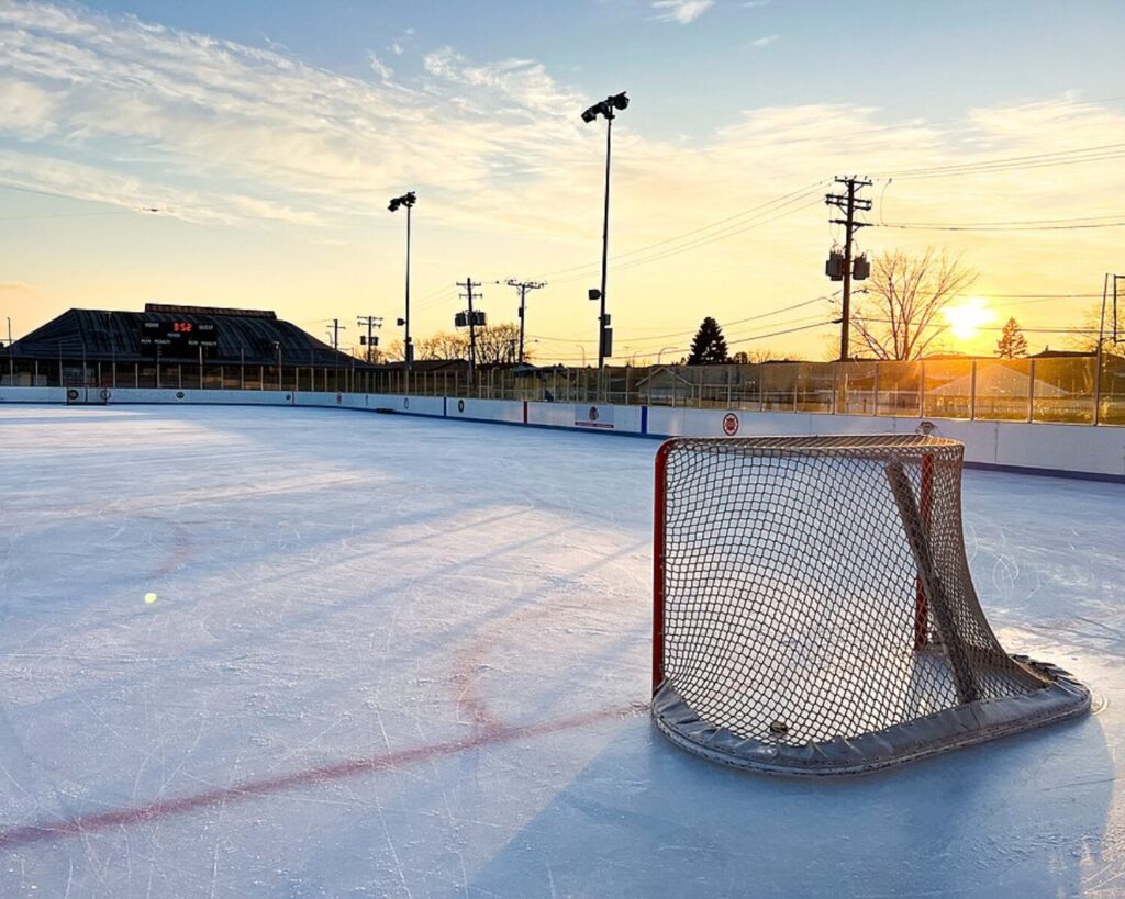Ice hockey rink at sunset with a red goal net in the foreground and empty boards along the sides.