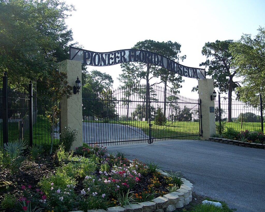 Dade City Pioneer Florida Museum entrance with gated archway, driveway, trees, and flower garden.