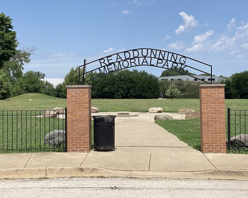 Read Dunning Memorial Park entrance with brick pillars, metal arch, fence, walkway, grass, and blue sky.