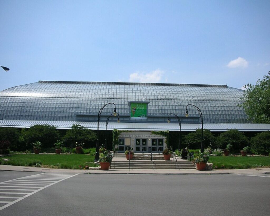 Garfield Park Conservatory exterior with glass greenhouse structure, entrance steps, gardens, road, and clear sky.