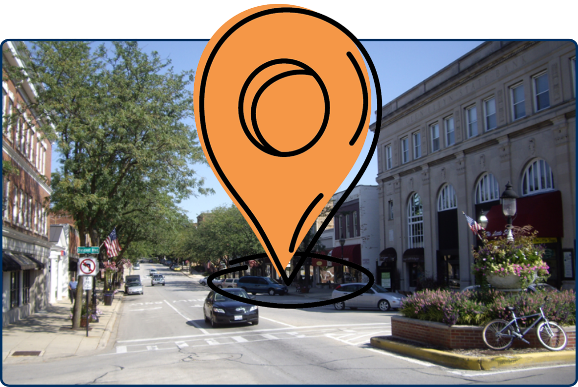 Downtown Glen Ellyn street lined with historic buildings, trees, shops, parked cars, and decorative planters under a clear blue sky.