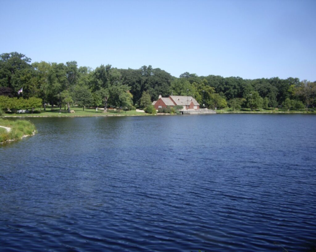 Lakeside park with a red brick building at the water's edge and a dock, surrounded by green trees under a clear blue sky