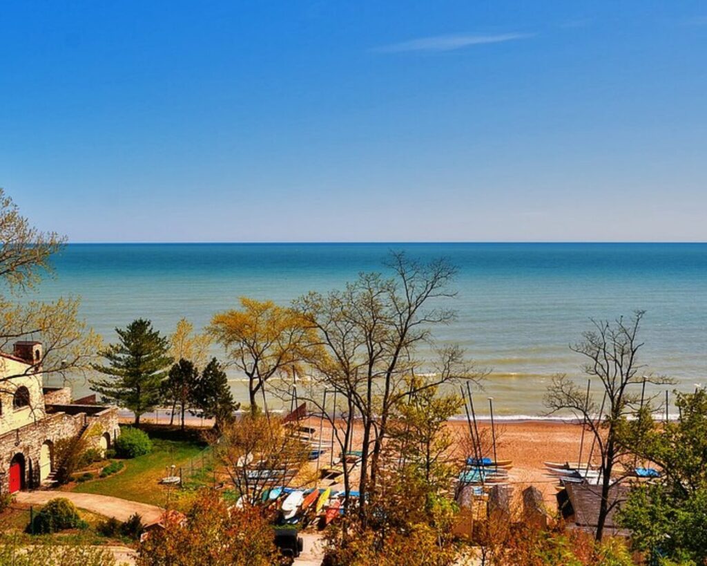 Elevated view of Glencoe’s Lake Michigan shoreline with a stone building, trees, beach, and small boats under a clear blue sky.