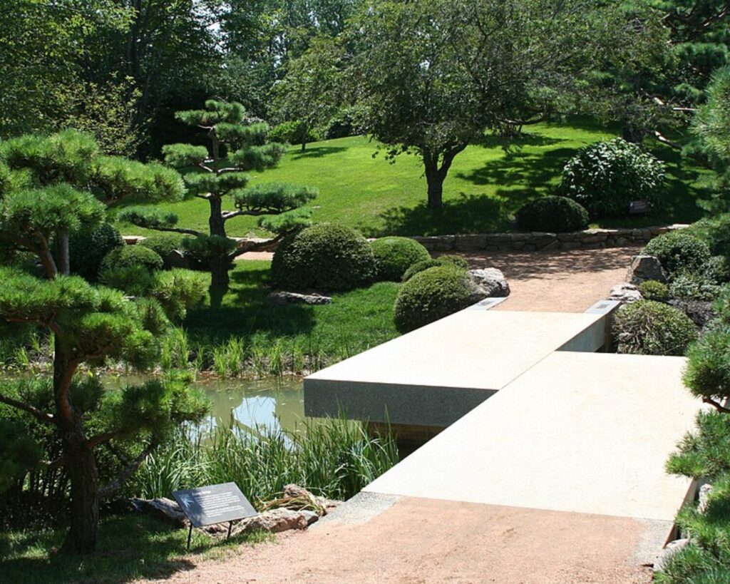 Tranquil Japanese-style garden with a rectangular concrete bridge over a pond, surrounded by manicured shrubs and green lawn
