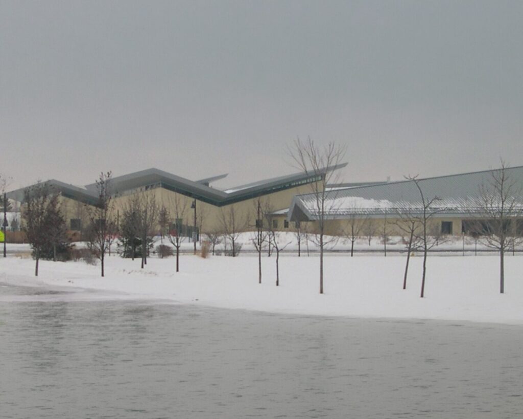 Snow-covered landscape with leafless trees in front of a modern beige building featuring angular, zigzag green roofs beside a frozen lake.