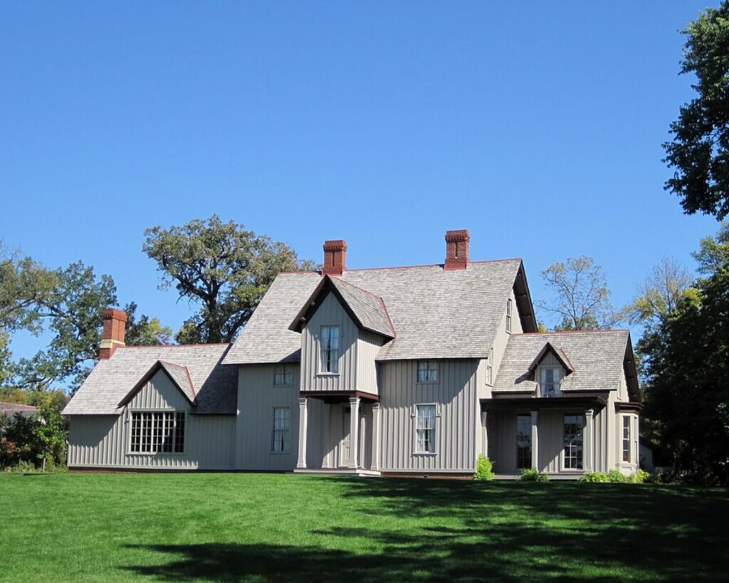Historic gray wooden house with steep gabled roofs and brick chimneys, set on a green lawn beneath a clear blue sky.