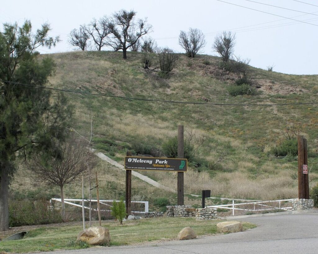 O’Melveny Park entrance with hillside, park sign, trees, fence, and cloudy sky.