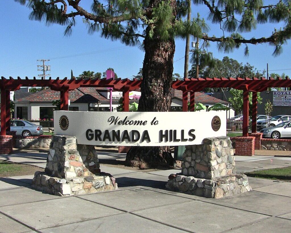 Granada Hills welcome sign with stone pillars, red pergola, large tree, sidewalk, and clear blue sky.