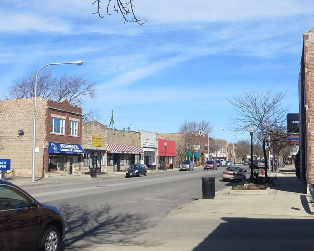 A sunny small-town street with brick storefronts and awnings, parked cars along the curb, and leafless trees against a blue sky.