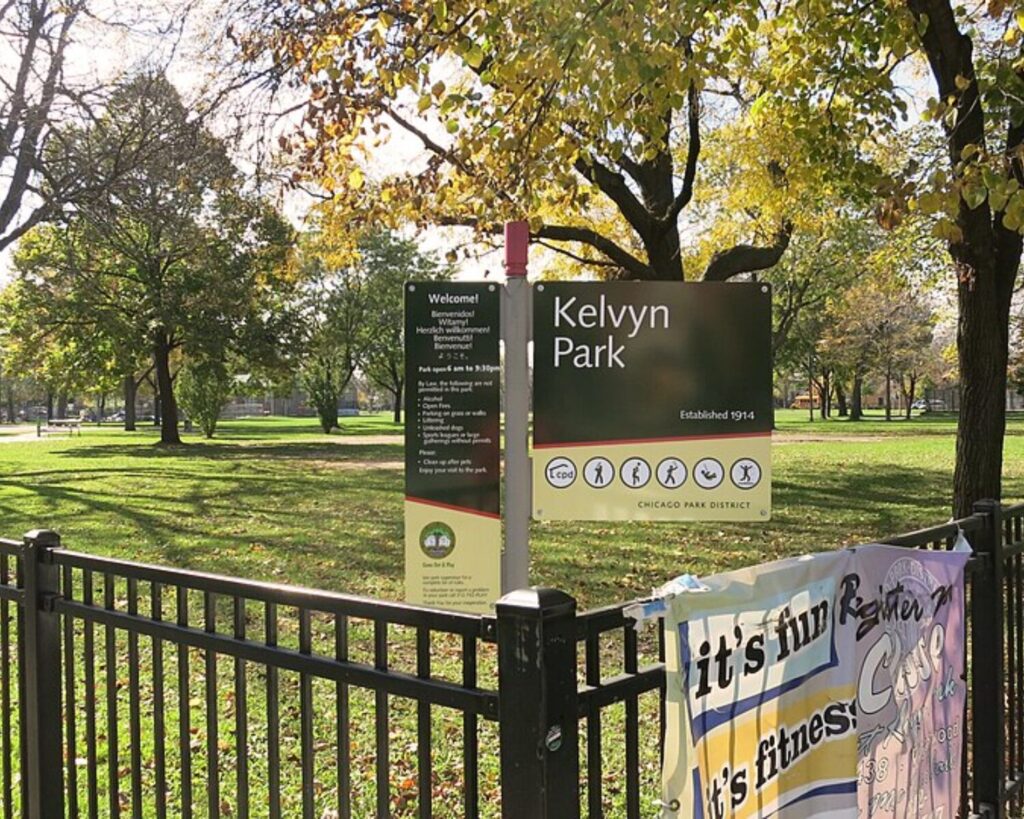 Kelvyn Park entrance sign beside a black fence, with a grassy lawn and mature trees in Chicago’s Hermosa neighborhood.