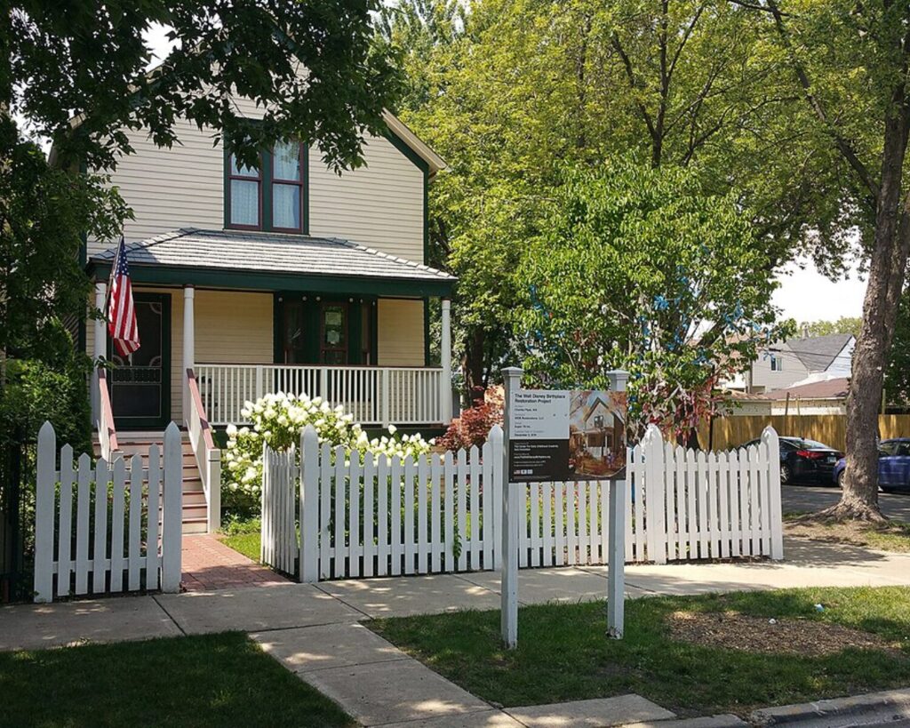 Two-story beige house with green trim, white picket fence, and a front porch; an American flag hangs by the steps and a sign stands in the yard about a restoration project.