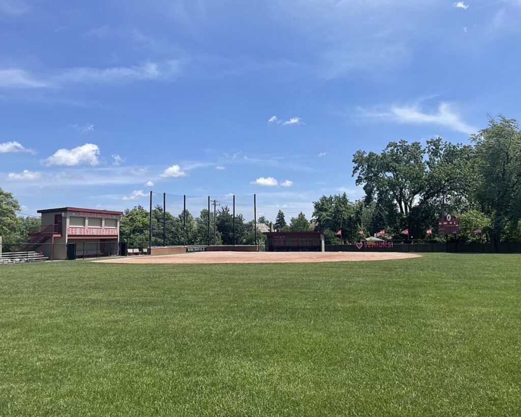 Softball field at Hinsdale Central High School with a green outfield, red dugouts, bleachers, and scoreboard beneath a bright blue sky.