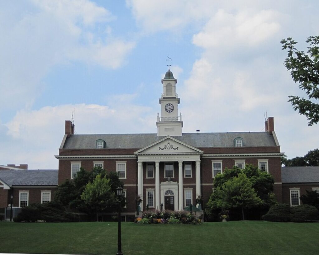 Brick campus building with white columns and a tall clock tower beneath a blue sky with clouds