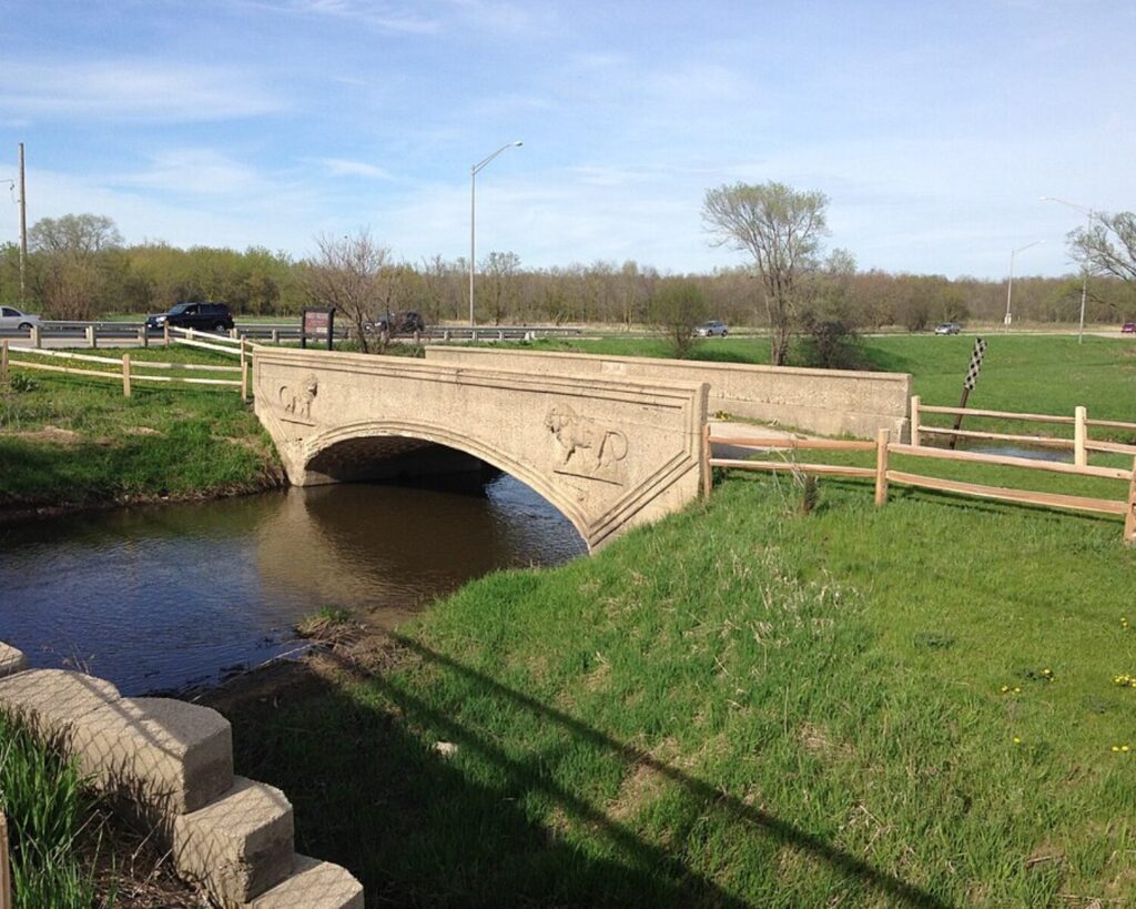 Historic concrete arch bridge crossing a small creek in a grassy Hoffman Estates landscape, with a roadway and cars in the background.