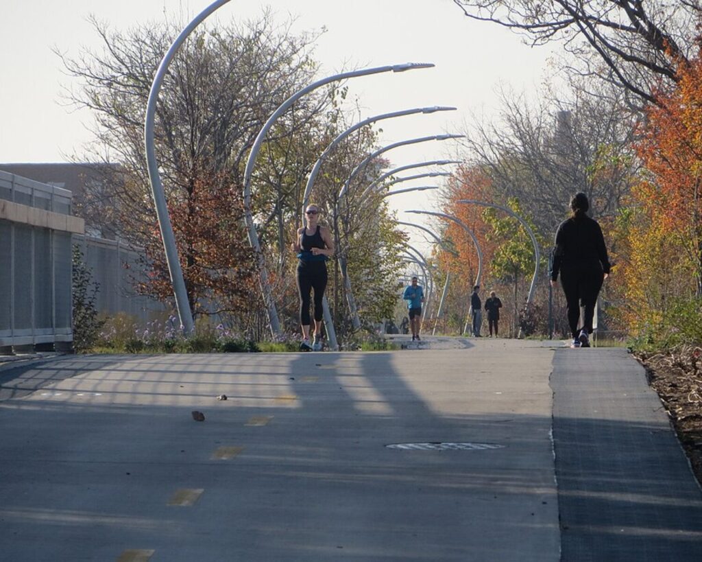 People jog and walk along a tree-lined paved path with curved metal arches overhead on an autumn day.