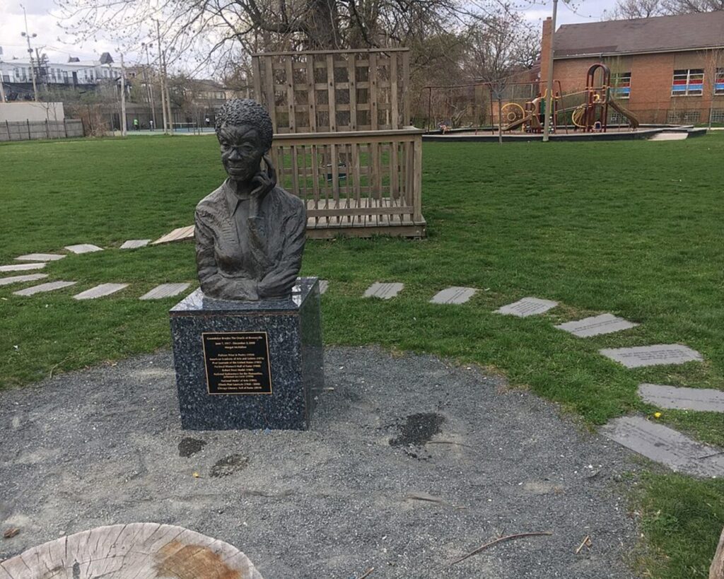 Bronze bust statue of a man on a speckled stone pedestal in a park with a playground in the background