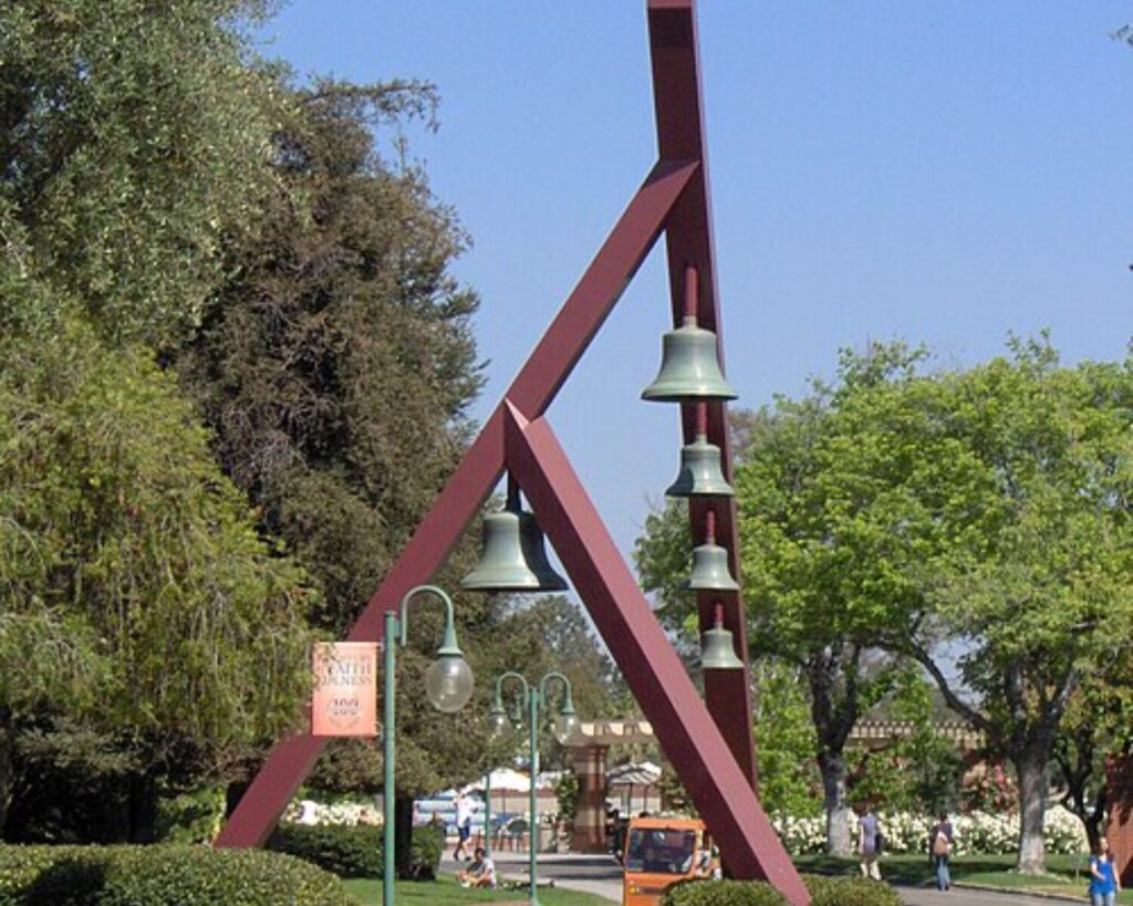 Large red triangular metal sculpture with hanging bells in a sunny park setting.